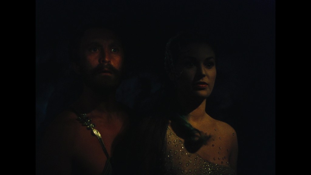 A dimly lit scene featuring a man with a beard and a woman in a sparkling dress, both looking forward with serious expressions.
