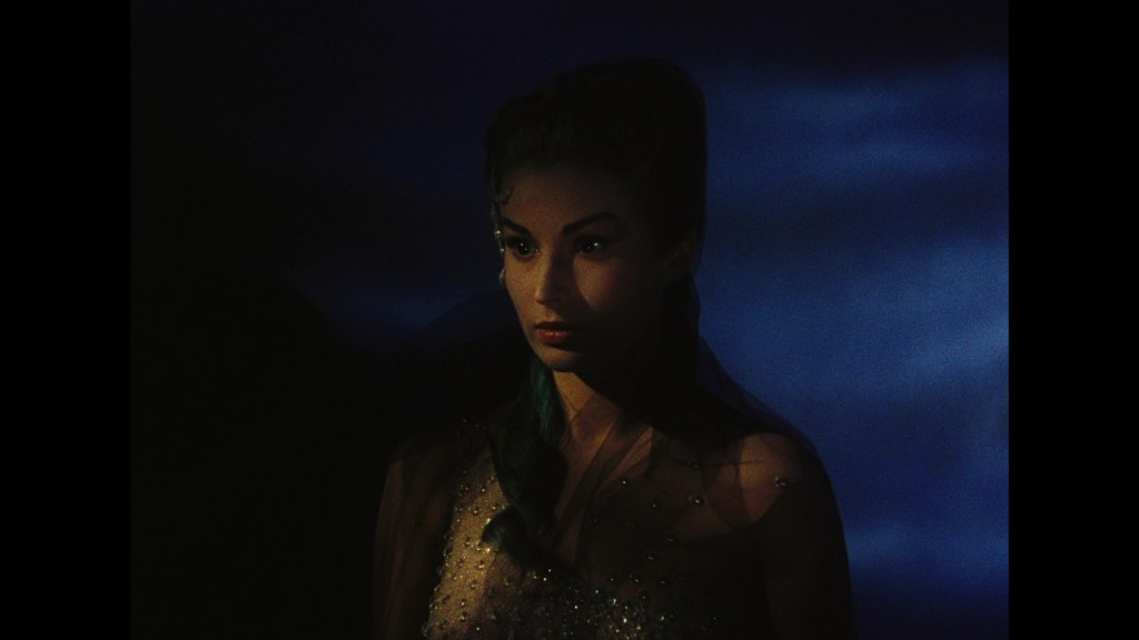 Close-up of a woman with dark hair and intense expression, wearing a sheer, beaded garment against a dark, moody background.
