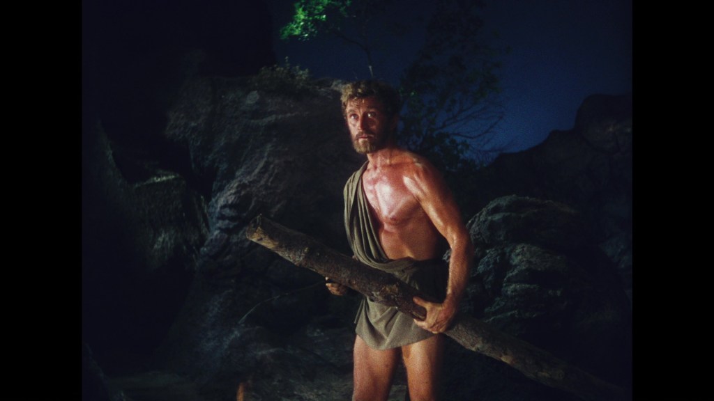A muscular man wearing a toga stands on rocky terrain at night, holding a large stick.
