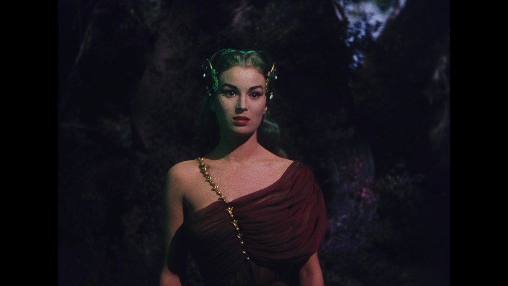 A woman with long hair and a dramatic expression, dressed in a flowing brown garment, stands in a dark, forest-like setting.