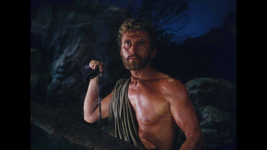 A muscular man with a beard, wearing a draped garment, stands in a rocky environment, holding a weapon, against a dark background.