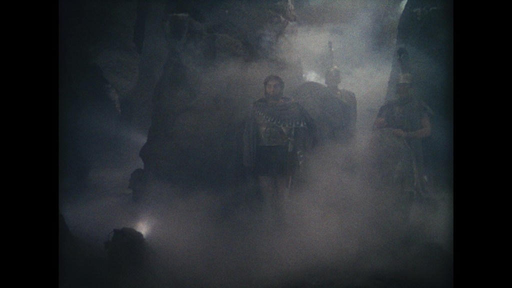 Three figures in ancient warrior attire stand amidst a foggy landscape, surrounded by dark rocky formations.