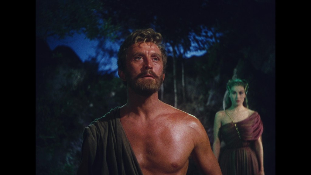 A muscular man with a beard stands in a forested area, looking upward with a serious expression. A woman in a purple dress stands behind him, partially visible.