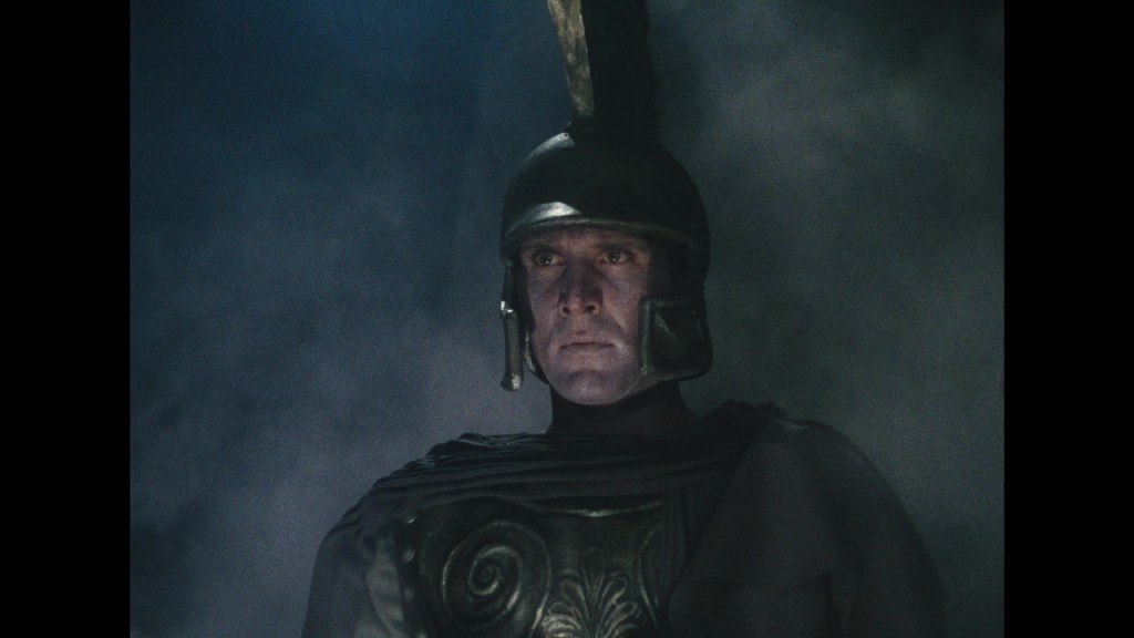 A solemn soldier wearing a traditional Roman helmet and armor, standing in a misty environment.