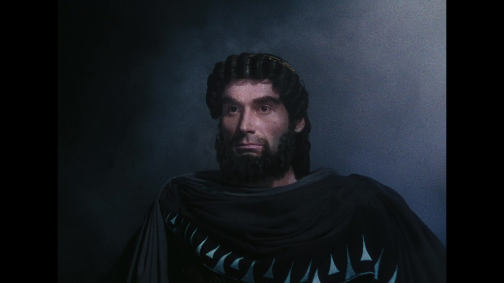 A dramatic close-up of a man with curly black hair and a beard, dressed in a dark cloak adorned with patterns, set against a smoky background.