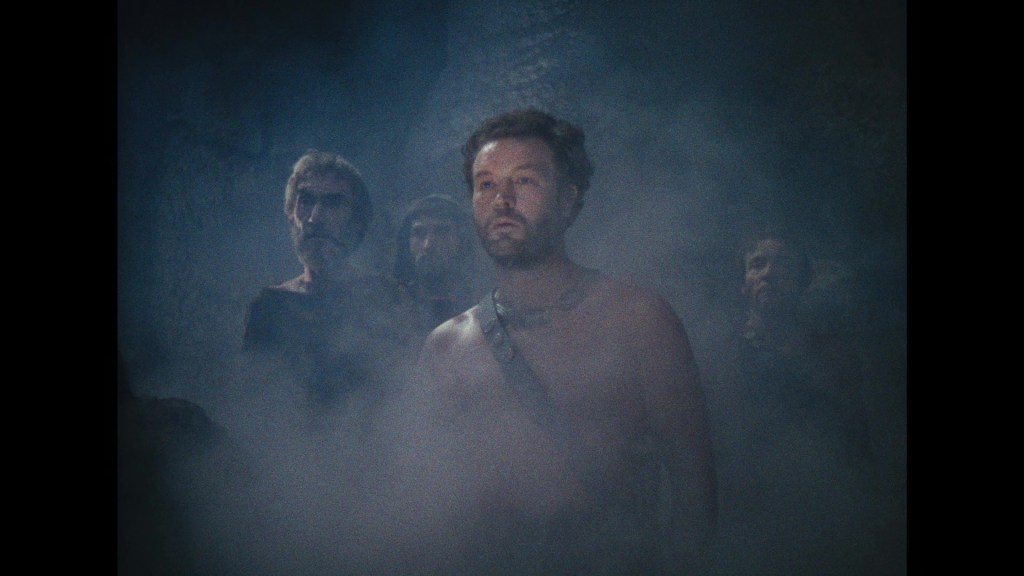 A group of ancient warriors stands in a foggy environment, with one man in the foreground appearing shirtless and looking serious, while others are partially obscured in the background.