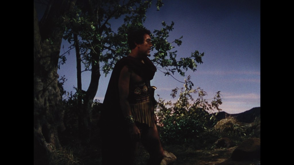 A figure dressed in ancient warrior attire stands in a natural setting, with greenery and a sky in the background.