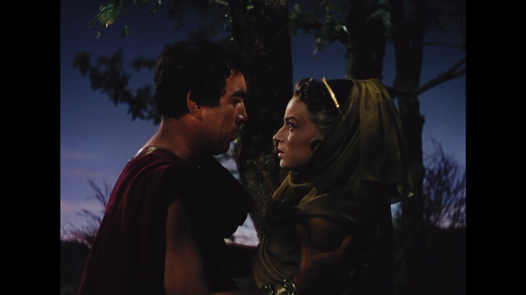 A dramatic scene featuring a man and woman standing close together, with intense expressions. The man is dressed in a toga-like garment, while the woman is draped in a flowing veil. They are surrounded by nature, with a dusk sky in the background.