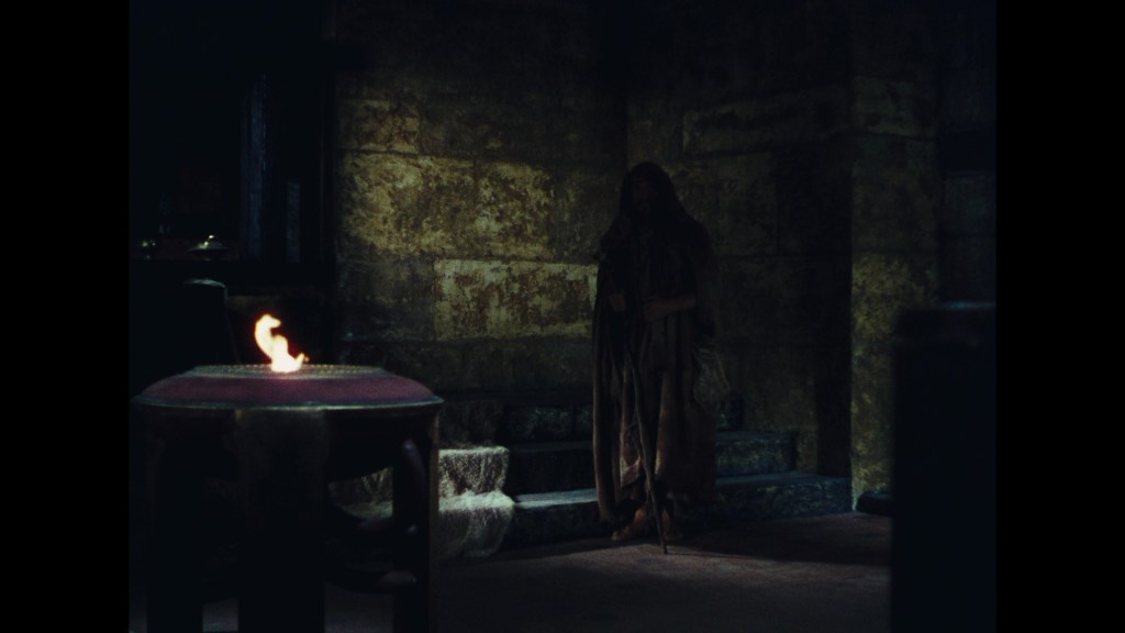 A dimly lit interior featuring a cloaked figure standing beside a table with a small flame, set against a backdrop of stone walls.
