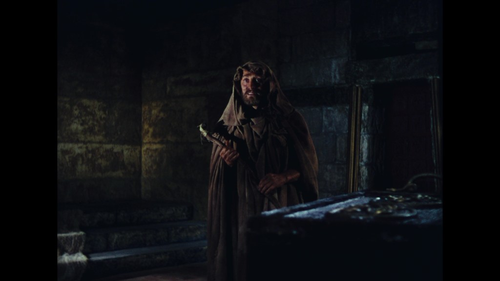 A cloaked figure stands in a dimly lit, ancient stone chamber holding a staff, with stone steps in the background.