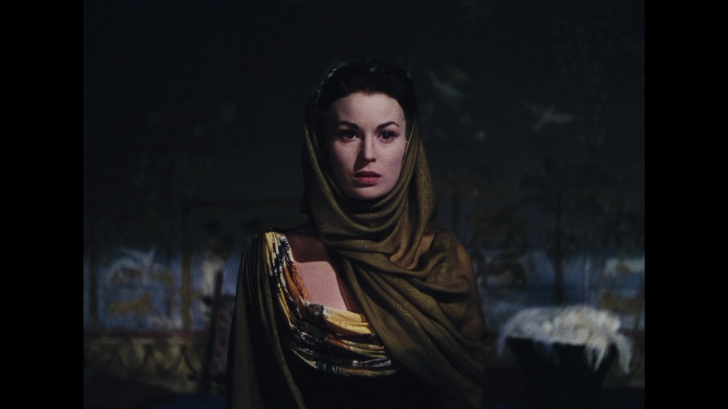 A woman in a dark setting wearing a gold and black patterned garment and a matching scarf, looking directly at the viewer with a serious expression.