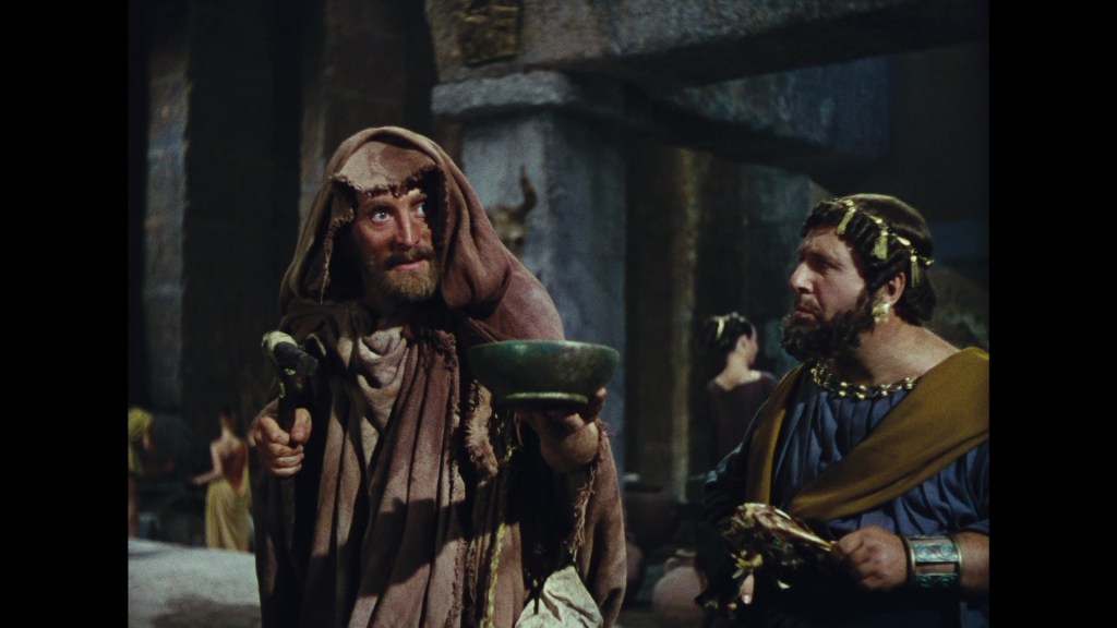 Two men in ancient costumes converse in a dimly lit setting, with one holding a bowl and a stick, while the other looks on, wearing a toga.
