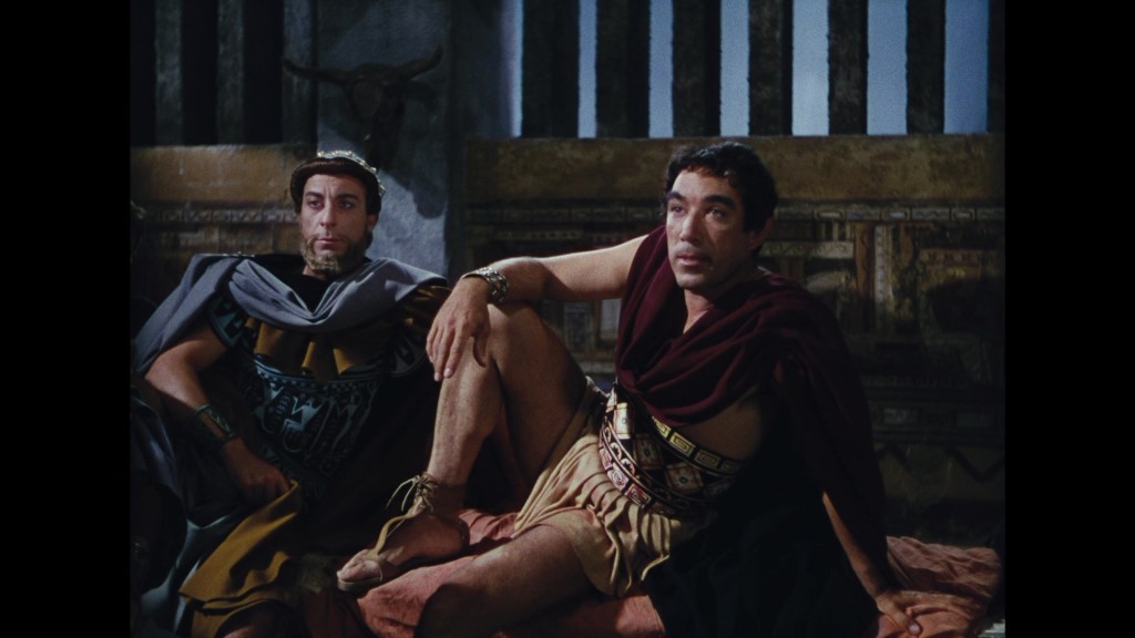 Two male figures in Roman-style clothing sitting on a couch, one with a contemplative expression and the other with an attentive posture, against a background of ancient decor.