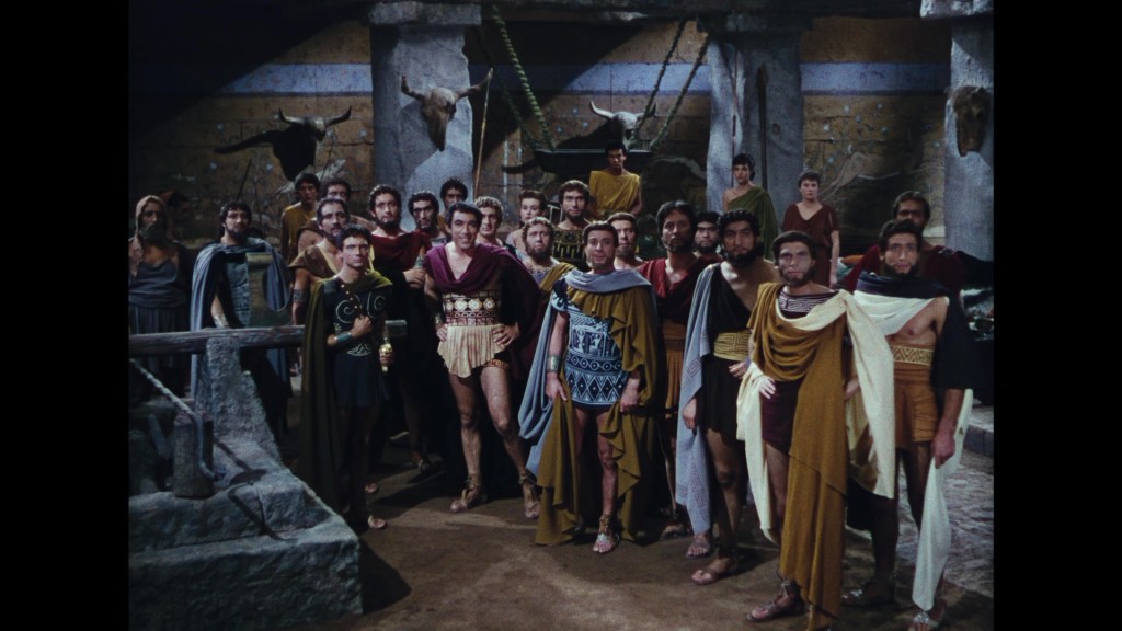 A group of men dressed in ancient Greek-style costumes standing in a stone chamber with animal horns mounted on the walls.