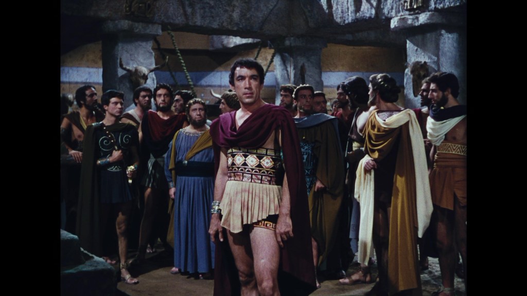 A group of men in ancient Greek attire, with one man in the foreground wearing a decorative tunic and a cape, standing confidently among others in a historical setting.