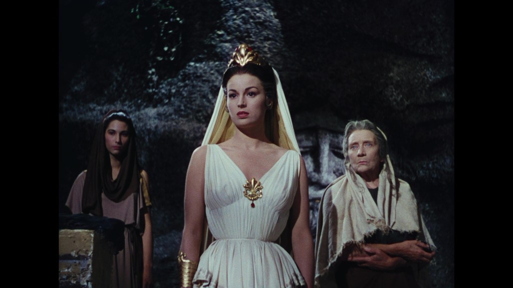 Three women in historical costumes stand in a dramatic setting. The central figure wears a white dress with gold accents and a crown, while the others wear muted tones and look on with varying expressions.