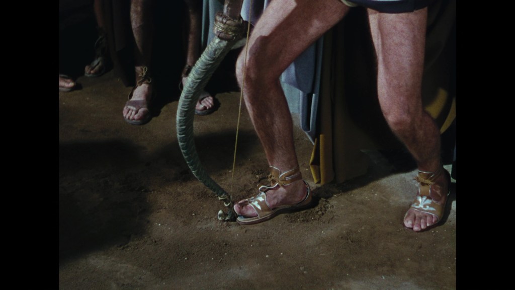 Close-up of a person's legs wearing sandals, standing on the ground with a rope or whip held in one hand, surrounded by others in the background.