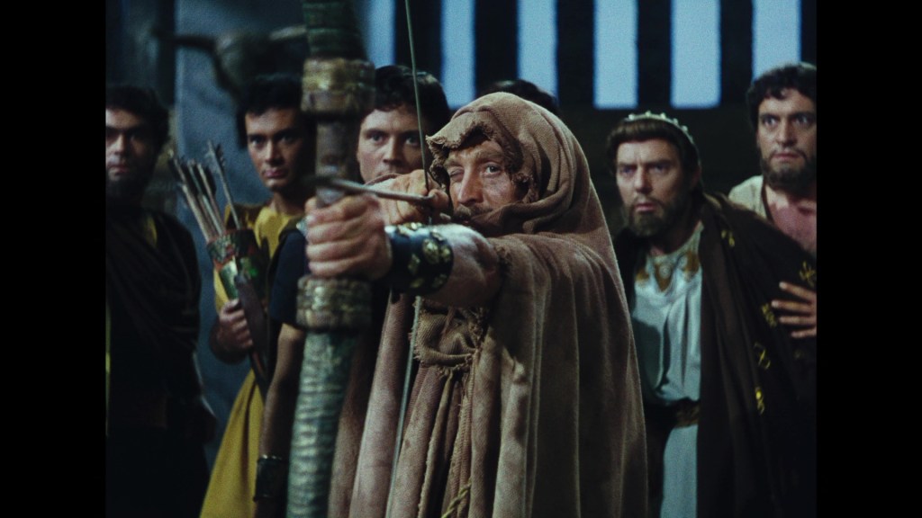 A group of men in historical attire, with one man in the foreground drawing a bow and arrow, while others watch intently.