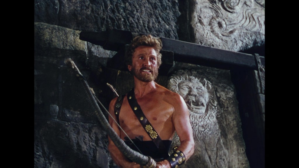 A muscular man with a beard stands shirtless, holding a bow, against a stone wall with intricate carvings.