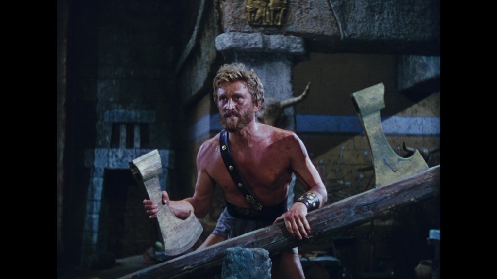 A muscular man with a beard and long hair, wearing minimal clothing, stands in a dark, ancient setting, gripping a large, wooden beam and preparing to lift a massive axe.