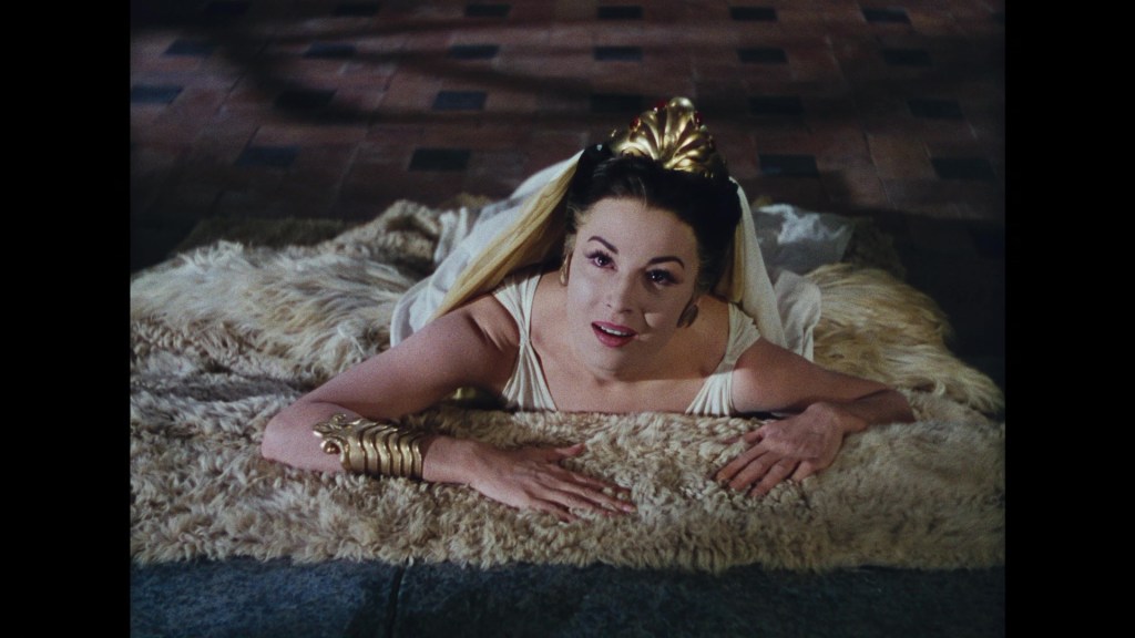 A woman lying on a fur rug, looking up with a surprised expression, wearing a dress and a decorative headpiece.