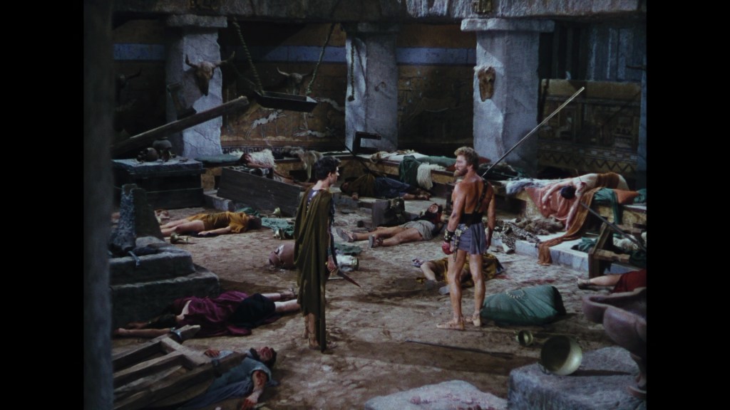 A dramatic indoor scene depicting two ancient warriors in conversation, surrounded by fallen bodies on the ground. The setting features stone walls, scattered debris, and remnants of battle.