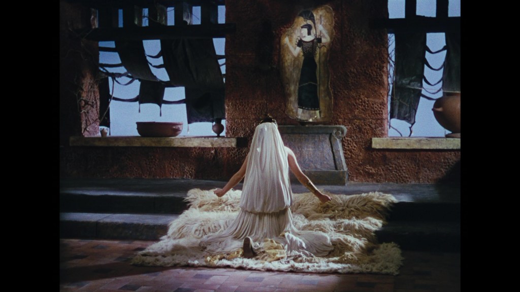 A woman in a flowing white dress kneels on a fur rug, facing a wall with a large painting of a woman in a black dress. The setting has a rustic, ancient feel with large windows and pottery visible in the background.