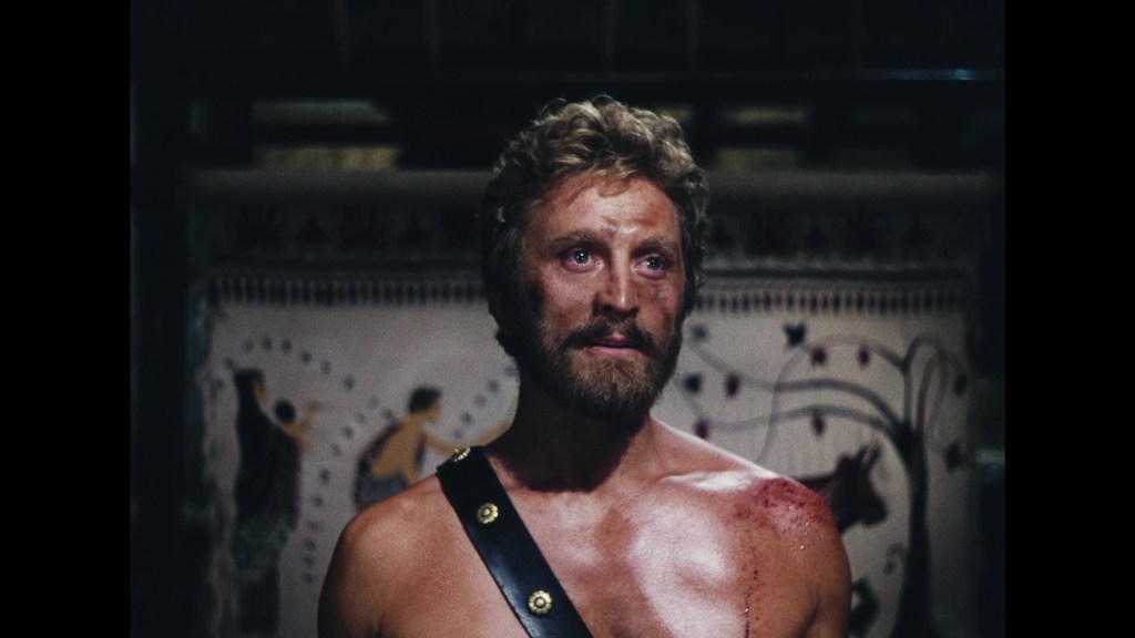 A rugged man with a beard and tousled hair, showing emotional expression, wearing a leather strap across his chest, with visible wounds on his shoulder, set against a backdrop featuring ancient artwork.