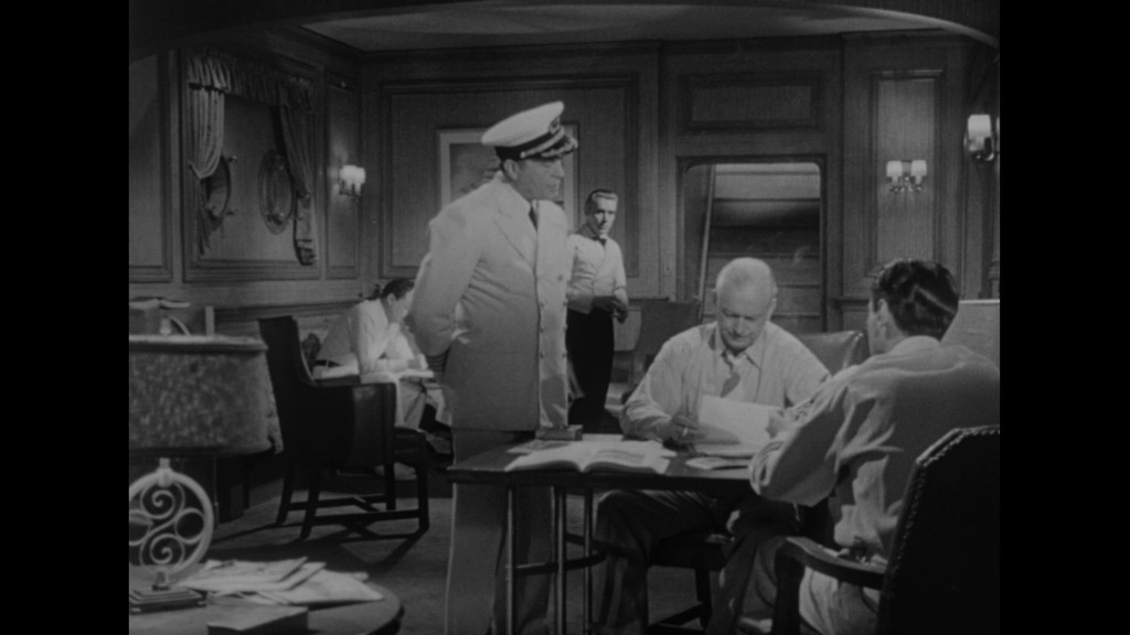 A black and white scene depicting several men in a ship's cabin. One man in a white uniform stands while others are seated at a table covered in documents. The room features elegant decor with lamps and curtains.
