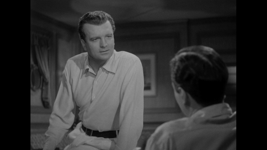 A black and white scene featuring a man in a light-colored shirt, looking thoughtfully while leaning against a table, having a conversation with another person whose back is partially visible.