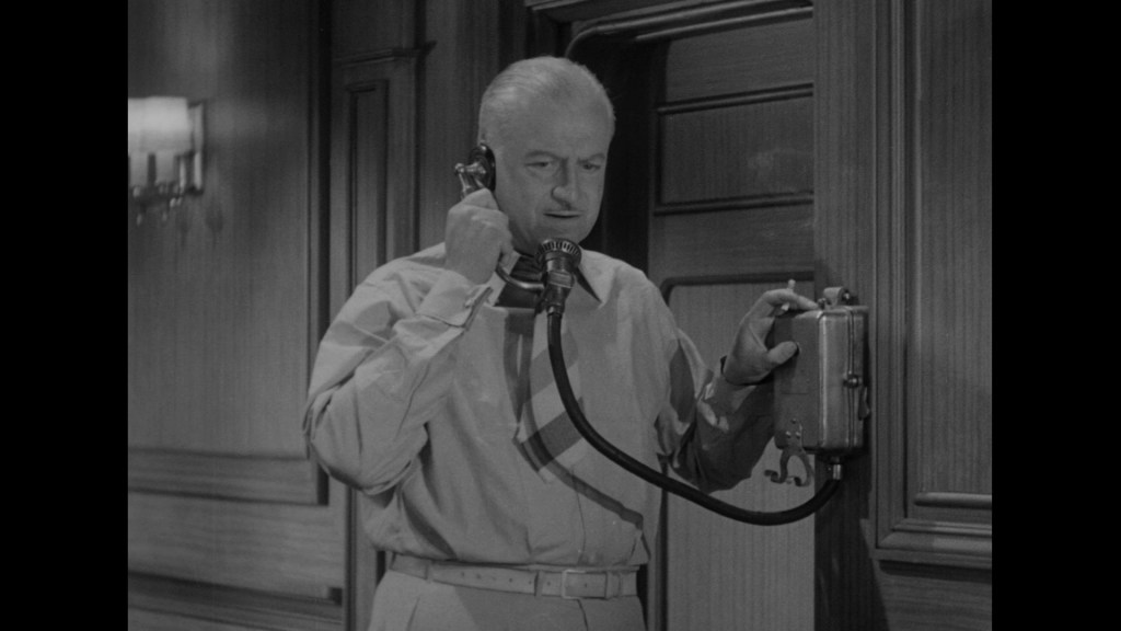 A man dressed in a button-up shirt is talking on a vintage telephone in a wood-paneled room.