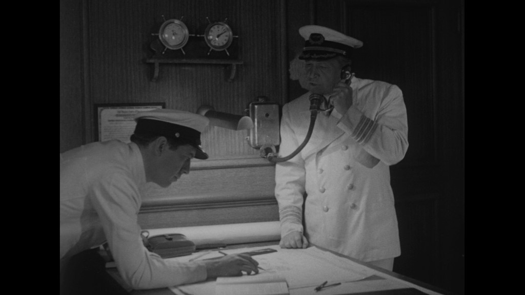 Two naval officers in a ship's control room; one stands talking on a telephone while the other studies a map on the table.