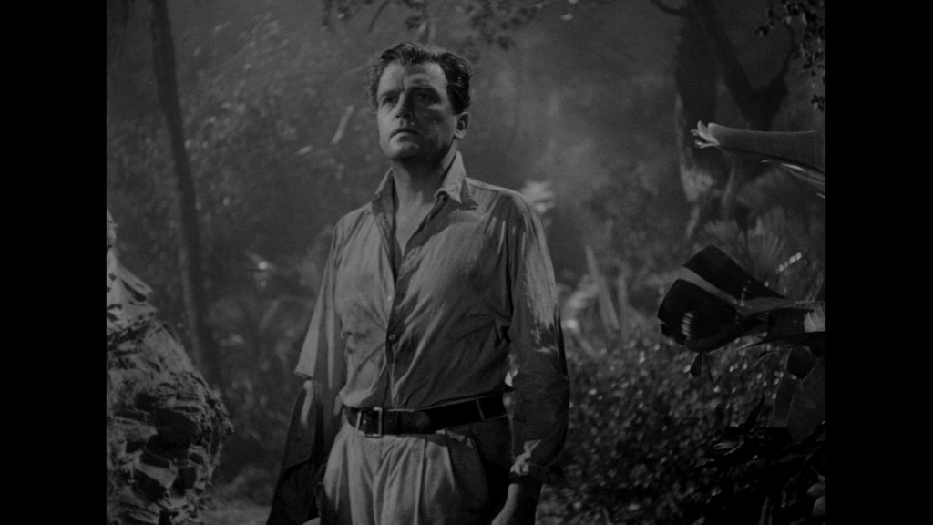 A man standing in a dark, forest-like environment, wearing a light-colored shirt and trousers, looking serious or contemplative.