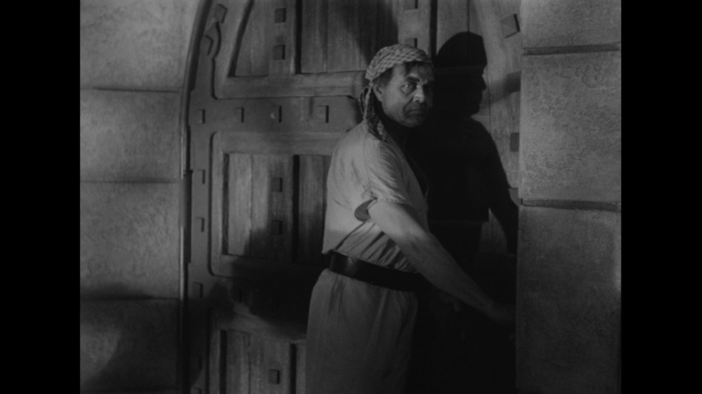 A man in a light tunic and headscarf stands at a large wooden door, appearing to be in a dark, stone setting. He has a serious expression and is partially turned away from the camera.
