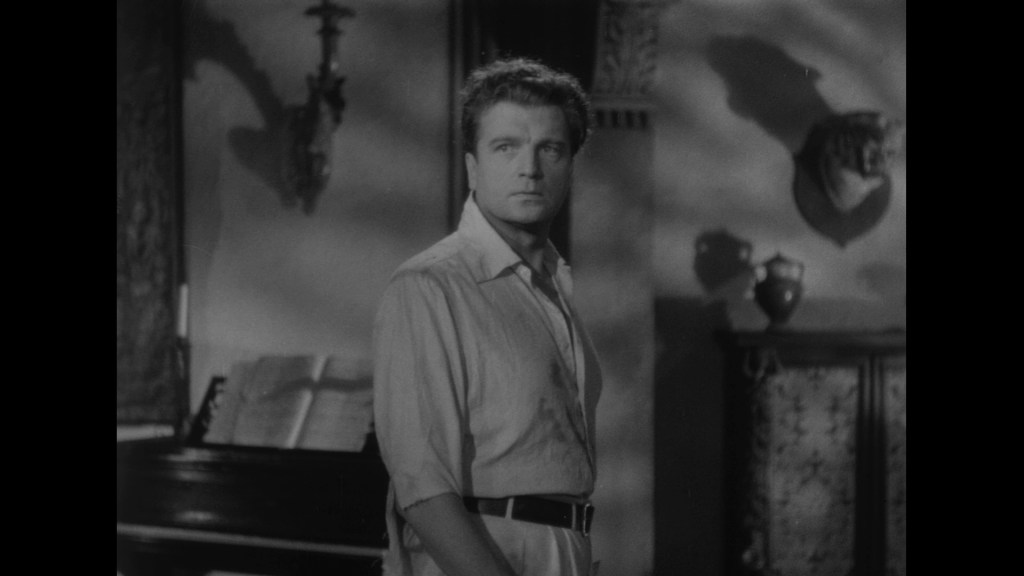 A man with tousled hair stands in a dimly lit room, looking back over his shoulder with a serious expression. The room features antique furnishings and a grand piano.