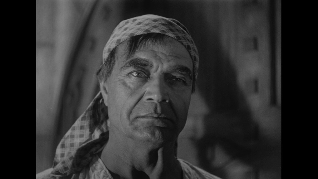 A close-up of an older man with a serious expression, wearing a scarf on his head, in a dimly lit interior setting.