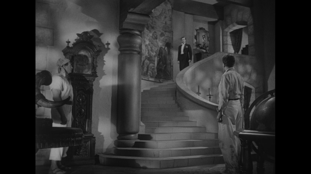 A dramatic black and white scene depicting two men standing in a room with a staircase, one man in formal attire descending the stairs while another in casual clothing looks up at him. The background features a large mural and ornate furnishings.