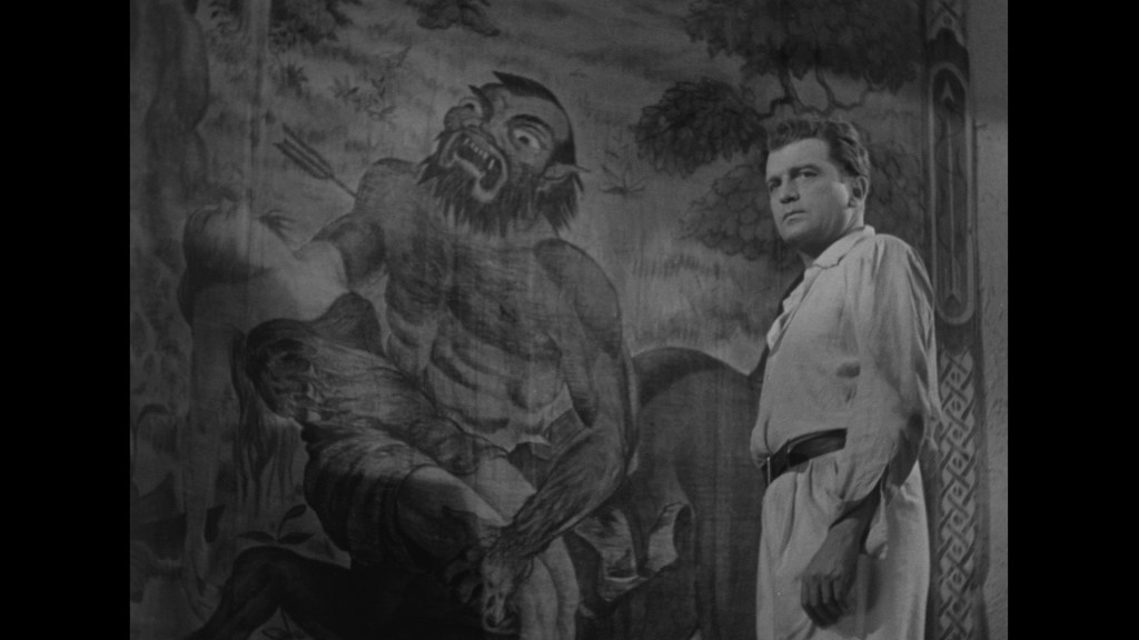 A man in a light-colored shirt stands in front of a large mural depicting a mythical figure with a fierce expression, surrounded by nature.