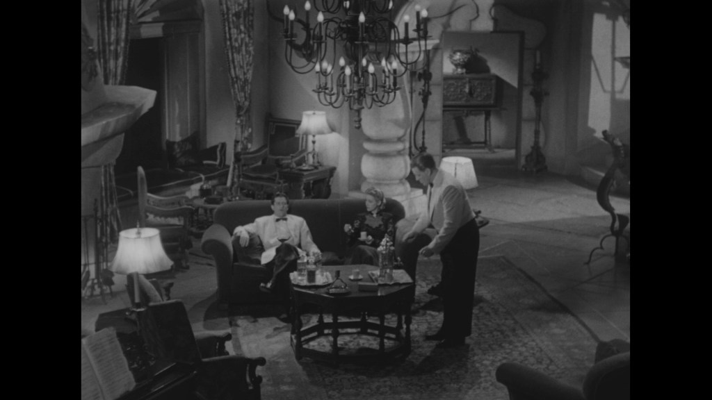 A vintage black and white scene depicting three people in a lavish living room, sitting on a couch and conversing over drinks, with elegant furnishings and a chandelier overhead.
