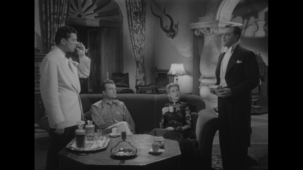 A group of four individuals engaged in conversation in a stylish, vintage setting. Two men, one in a white suit and the other in a black tuxedo, stand while a man in casual attire and a woman in a black dress sit on a couch. The scene features elegant furnishings and decorative elements typical of classic cinema.
