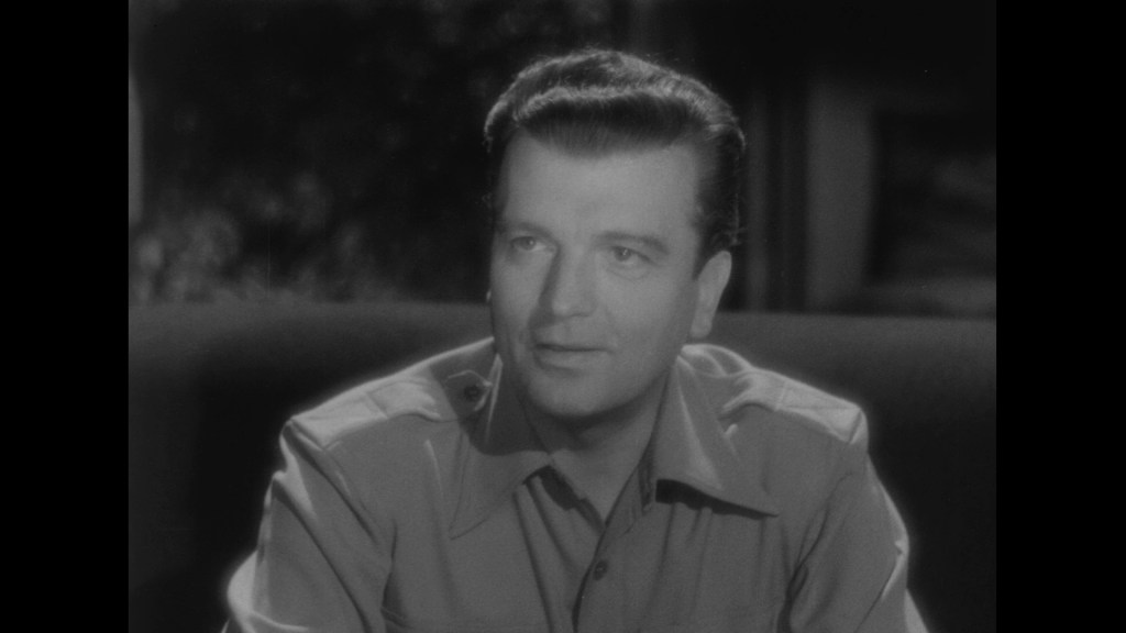 A man in a button-up shirt sitting on a couch, looking thoughtfully to the side in a black and white setting.