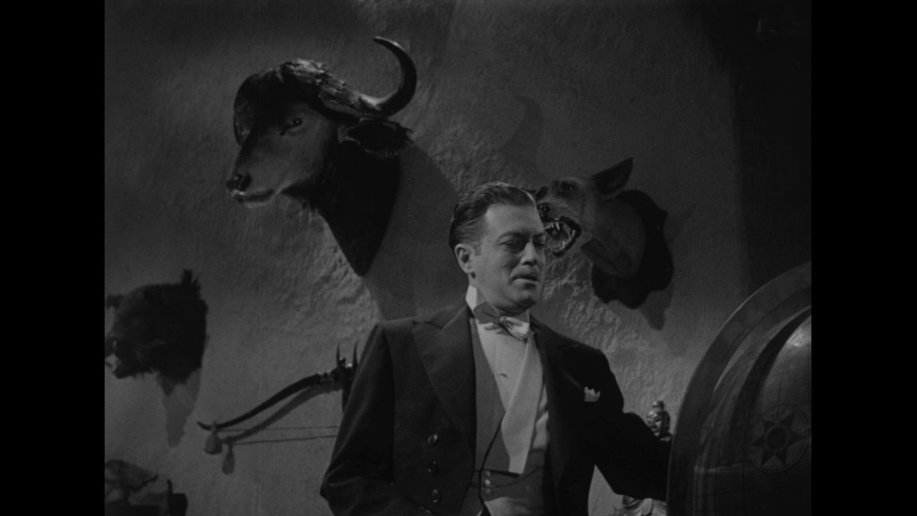 A man in a tuxedo stands in a dimly lit room with animal heads mounted on the walls, including a bull and a wolf. The atmosphere is dark and moody.