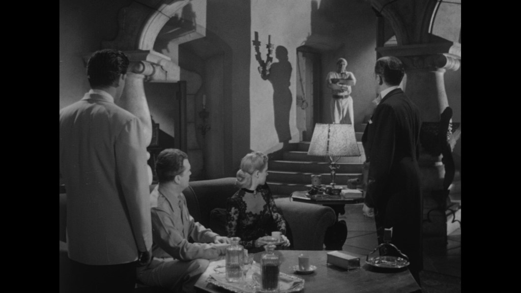 A group of four people, dressed in 1940s attire, are gathered in a vintage living room setting, with two men standing and one woman seated. A figure in the background, partially obscured in shadow, stands by a doorway holding a mysterious object. The atmosphere is tense, and dim lighting casts shadows across the scene.