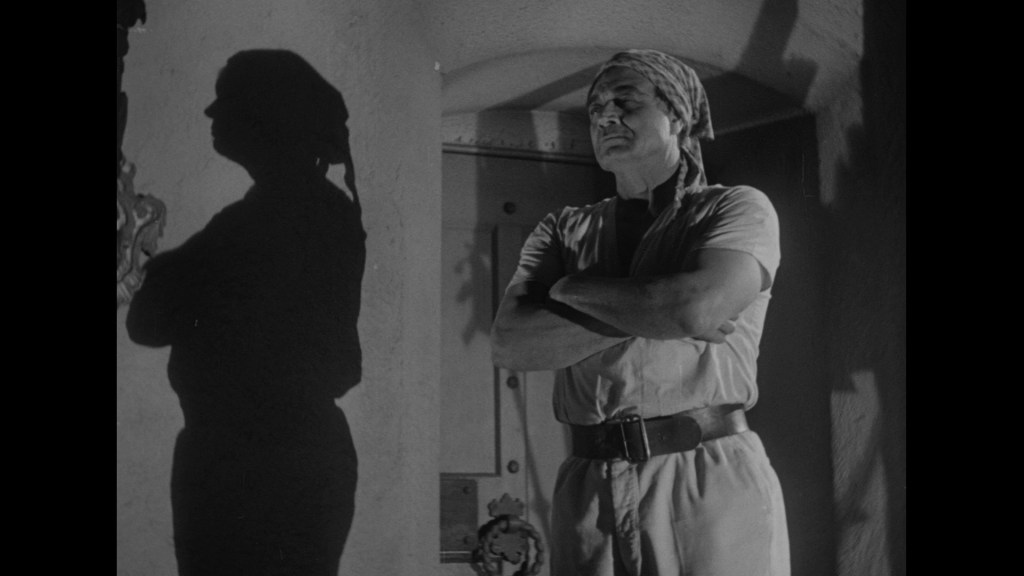 A man stands with his arms crossed, wearing a headscarf and a light-colored outfit, cast in shadow against a textured wall.