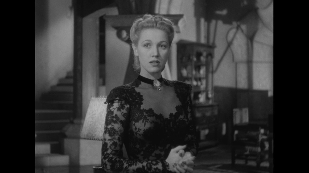 A woman in a black lace dress, standing in an interior setting with stairs and furniture in the background.