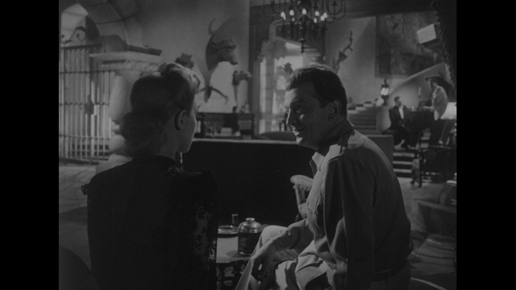 A scene in a vintage black and white setting featuring a man and a woman sitting close together, engaging in conversation. The background showcases a stylish, dimly lit room with decorative elements, including a staircase and various artistic pieces.