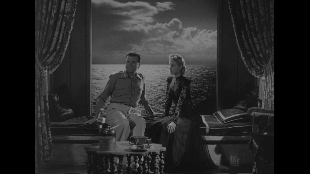 A man in military attire and a woman in a vintage black dress sit on a window seat, overlooking a serene ocean view with a cloudy sky in the background.