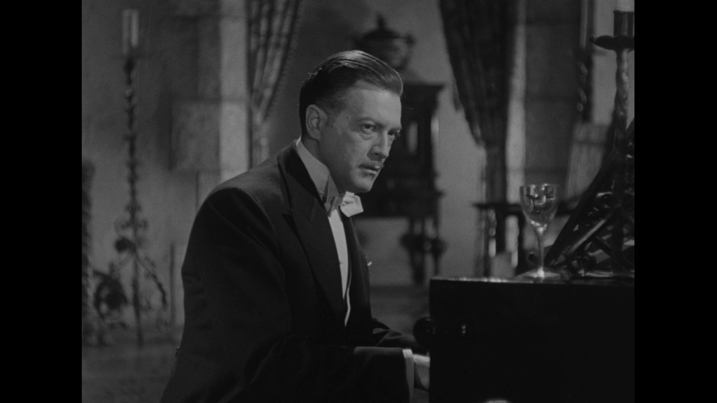 A man in a tuxedo sitting at a grand piano in a dimly lit room, with a glass placed nearby.