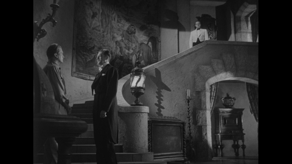 A dimly lit grand staircase in a mansion, featuring two men in formal wear standing face to face, with one man on the stairs and the other at the bottom. A figure in white stands above them on the landing, silhouetted against a large mural.