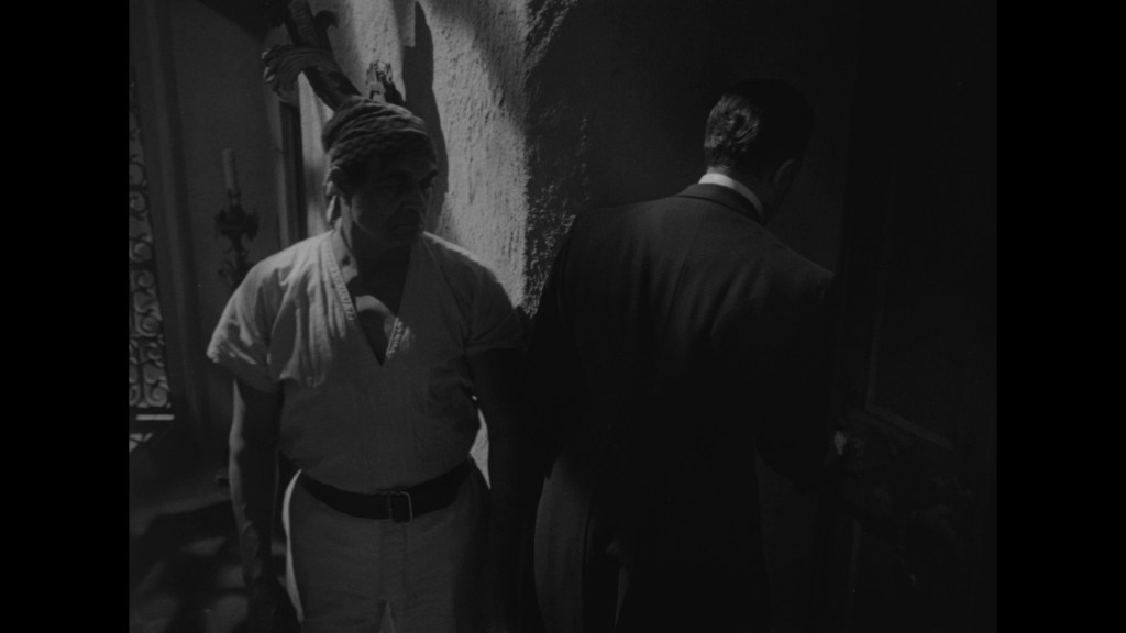 A shadowy black and white scene featuring a man in a loose white shirt standing beside another man in a dark suit, who is partially turned away as he approaches a door.